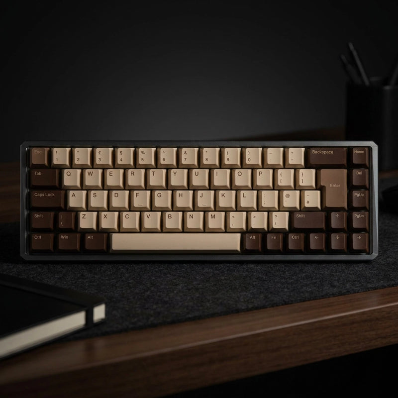 A compact mechanical keyboard with a dark gray frame and beige and brown keycaps, resting on a dark desk mat on a wooden desk.