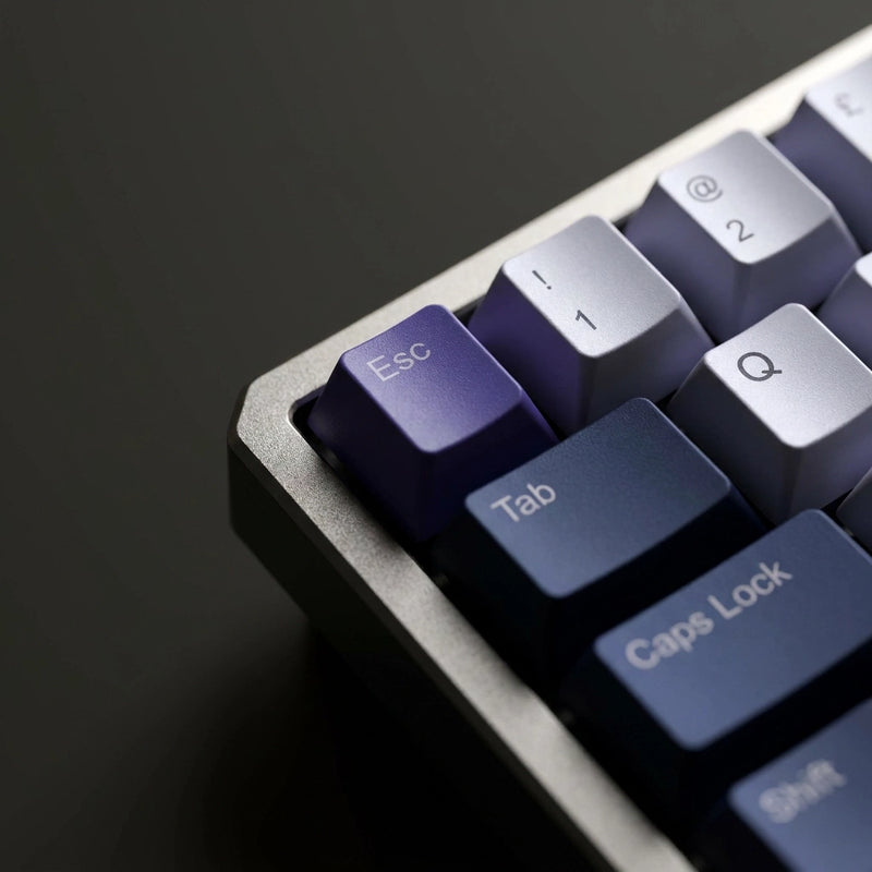Close-up of a mechanical keyboard showing keycaps in a gradient from purple to dark blue, set against a dark gray background.