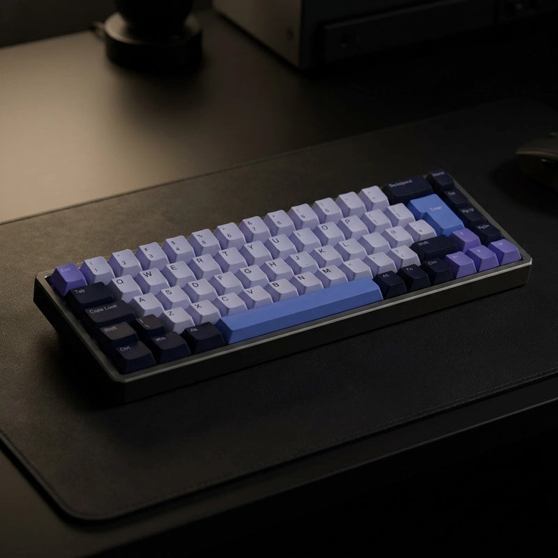 A mechanical keyboard with a custom keycap set in shades of light blue, lavender, and dark navy, resting on a black desk mat.
