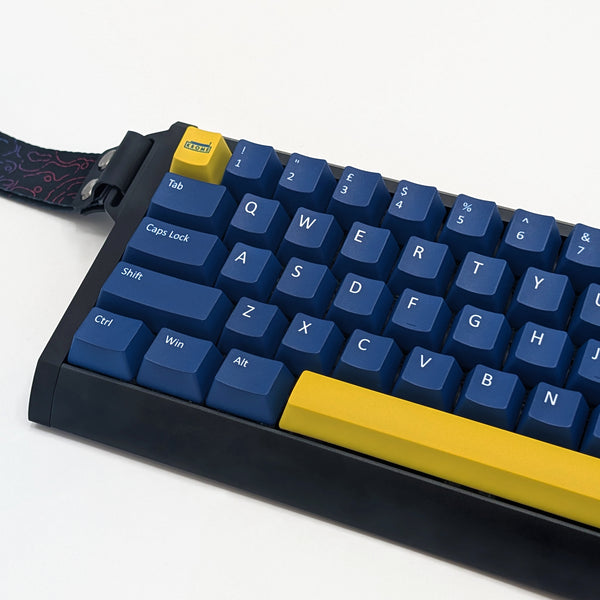 A close-up of a mechanical keyboard with blue keycaps, a black frame, and bright yellow accent keys, including a large bottom-right modifier key and a small top-left tab key. A patterned wrist strap is attached on the left.