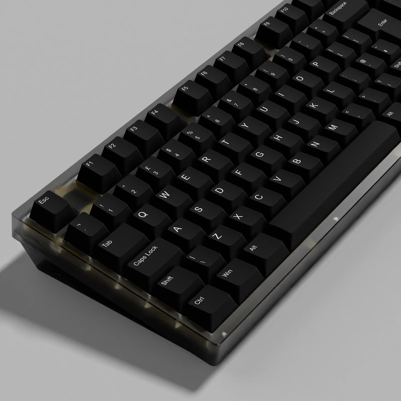 A close-up of a mechanical keyboard with black keycaps and a translucent dark gray case, shown at an angle on a neutral gray background.